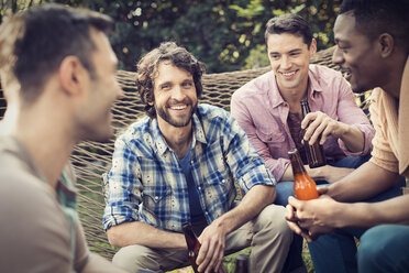 Eine Gruppe von Freunden liegt in einer großen Hängematte im Garten und trinkt ein Bier. - MINF03459