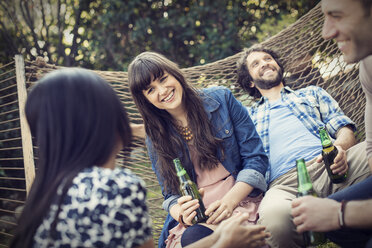 Eine Gruppe von Freunden liegt in einer großen Hängematte im Garten und trinkt ein Bier. - MINF03463