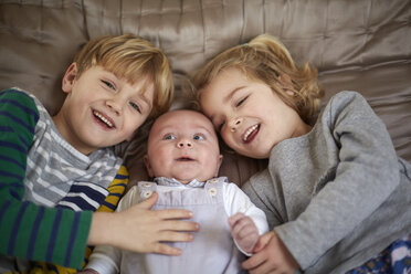 Three children lying on a bed, a boy and girl with a baby between them. - MINF03495