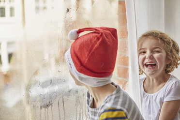 Ein Junge mit einer Weihnachtsmannmütze, der aus dem Fenster seines Zimmers schaut, und seine Schwester, die neben ihm lacht. - MINF03498