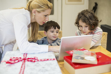 Zwei Kinder und ihre Mutter sitzen am Küchentisch und schauen auf einen Laptop-Bildschirm. - MINF03504