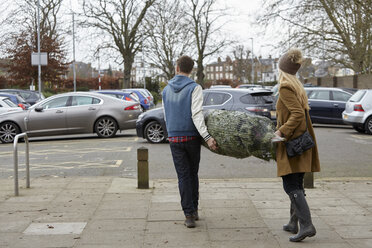 Zwei Personen tragen einen Weihnachtsbaum mit Netz zum Parkplatz. - MINF03508