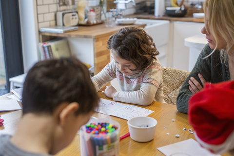 Drei Kinder und eine erwachsene Frau an einem Tisch, die Karten und Briefe an den Weihnachtsmann malen und schreiben., lizenzfreies Stockfoto