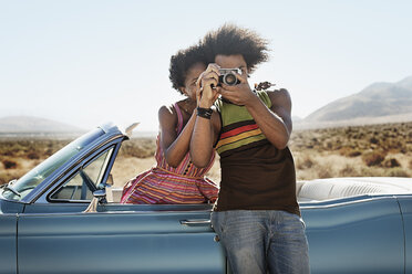 Ein junges Paar, Mann und Frau, in einem hellblauen Cabrio auf offener Straße, mit einer Kamera in der Hand. - MINF03588