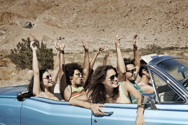 Eine Gruppe von Freunden in einem hellblauen Cabrio auf offener Straße, die über eine trockene, flache Ebene fahren, die von Bergen umgeben ist. - MINF03593