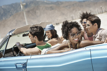 Eine Gruppe von Freunden in einem hellblauen Cabrio auf offener Straße, die über eine trockene, flache Ebene fahren, die von Bergen umgeben ist. - MINF03597