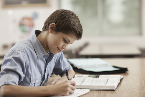 Ein Junge sitzt in der Klasse an einem Schreibtisch, benutzt Schulbücher und hält einen Stift. - MINF03652