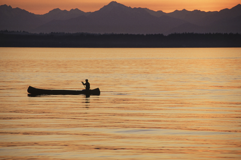 Eine Person sitzt in einem großen Kanu im indianischen Stil und paddelt bei Sonnenuntergang über ruhiges Wasser., lizenzfreies Stockfoto