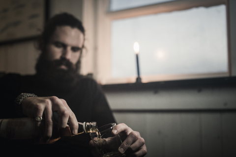 Ein Mann sitzt allein in einem Zimmer und schenkt sich ein Glas Whisky ein. Eine brennende Kerze., lizenzfreies Stockfoto