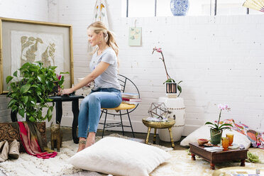 Eine junge Frau sitzt auf einem Stuhl in einer mit Bildern und Pflanzen dekorierten Wohnung. - MINF03675