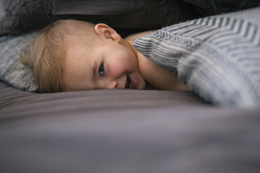 Ein kleiner Junge liegt auf dem Bauch im Bett, schaut in die Kamera und lacht. - MINF03684
