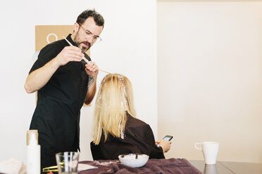 Ein Haarkolorist arbeitet mit Folien, um einer Kundin mit langem blondem Haar Strähnchen und Lichter mit Farbe zu geben. - MINF03744