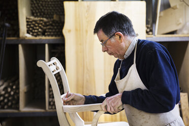 Ein Mann steht in einer Tischlerwerkstatt und arbeitet an einem Holzstuhl, wobei er das Gelenk der Armlehne mit einem Bleistift markiert. - MINF03746