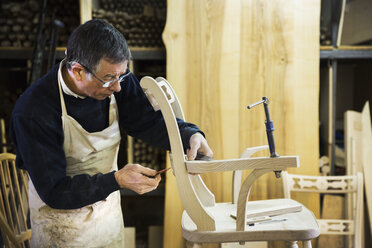 Ein Mann steht in einer Tischlerwerkstatt und arbeitet an einem Holzstuhl, wobei er das Gelenk der Armlehne mit einem Bleistift markiert. - MINF03747