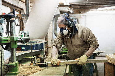 Mann in einer Tischlerwerkstatt, mit Atemschutzmaske und Gehörschutz, bei der Arbeit an einem Stück Holz. - MINF03751