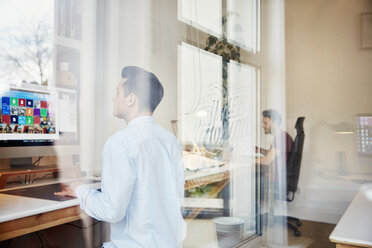 Zwei Männer arbeiten an Computern in einem Designstudio, Blick durch ein Fenster und Reflexionen des Fensterrahmens und der Bäume. - MINF03791