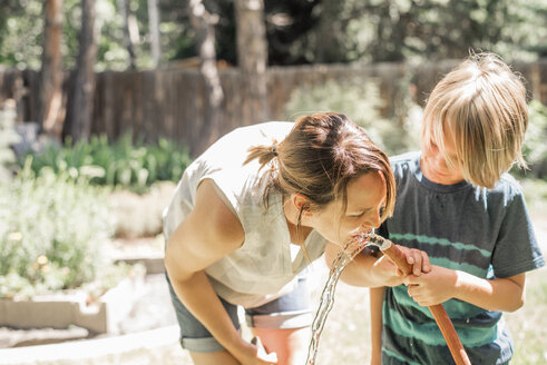 Mutter und Sohn stehen in einem Garten und trinken Wasser aus einem Gartenschlauch. - MINF03824