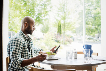 Mann mit kariertem Hemd sitzt in einem Café und benutzt sein Mobiltelefon. - MINF03850