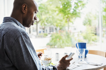 Mann mit kariertem Hemd sitzt in einem Café und benutzt sein Mobiltelefon. - MINF03851