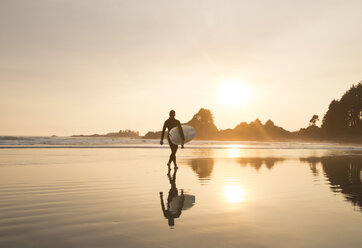 Reflection of man wearing wet suit and carrying surfboard walking along sandy beach at sunset. - MINF03916