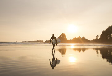 Spiegelung eines Mannes mit Neoprenanzug und Surfbrett, der bei Sonnenuntergang am Sandstrand spazieren geht. - MINF03916