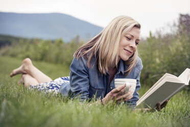 Eine Frau liegt im Gras, hält eine Teetasse und liest ein Buch. - MINF04115