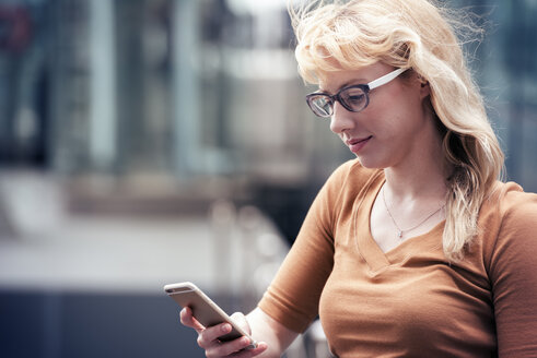 Eine blonde Frau auf einer Straße, die ihr Handy benutzt und nach Nachrichten sucht - MINF04149