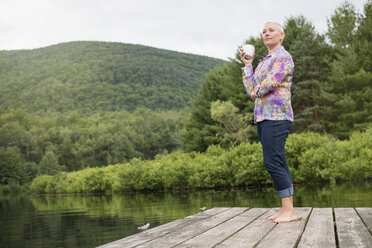 Eine Frau steht mit einer Kaffeetasse in der Hand auf einem Steg am See. - MINF04177