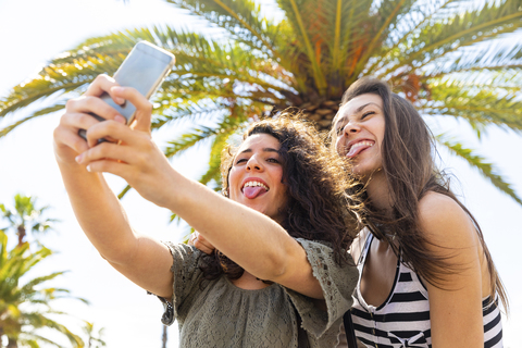 Two playful female friends taking a selfie under a palm tree stock photo