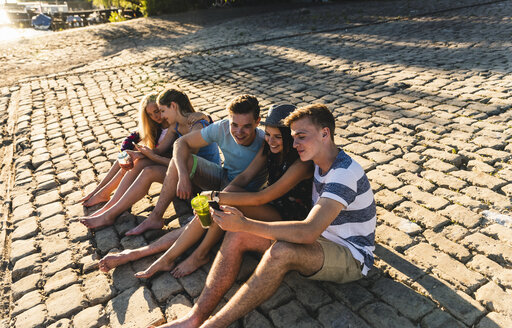 Eine Gruppe von Freunden sitzt mit Erfrischungsgetränken und Mobiltelefonen auf Kopfsteinpflaster - UUF14809