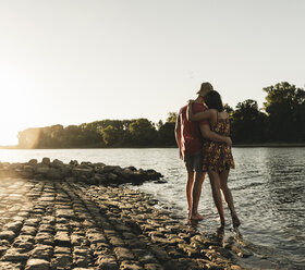 Rear view of young couple embracing at a river at sunset - UUF14820