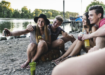 Eine Gruppe von Freunden sitzt zusammen, grillt und macht ein Selfie am Flussufer - UUF14841