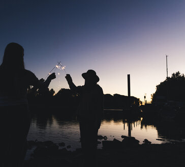 Silhouette von Menschen am Flussufer mit Wunderkerzen am Abend - UUF14857