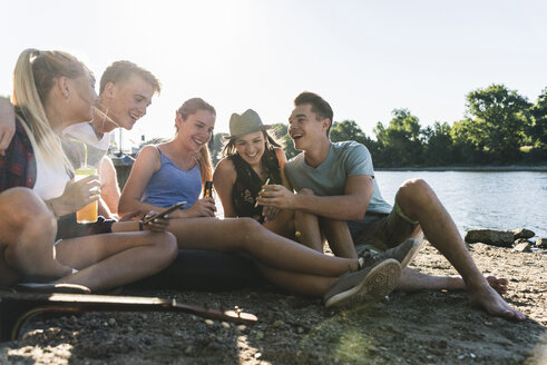 Eine Gruppe glücklicher Freunde sitzt am Flussufer mit Getränken und Mobiltelefon - UUF14903