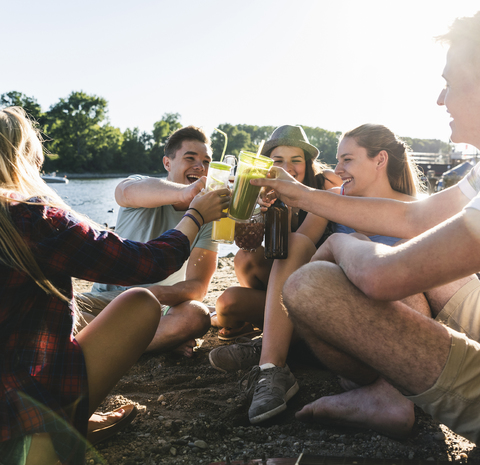Eine Gruppe glücklicher Freunde sitzt am Flussufer und stößt mit Getränken an, lizenzfreies Stockfoto