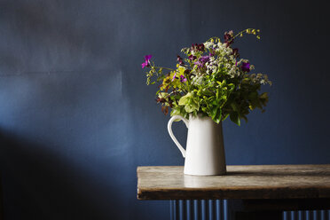 Nahaufnahme einer weißen Kanne mit einem Strauß Wildblumen auf einem Holztisch, dahinter eine blaue Wand. - MINF04399