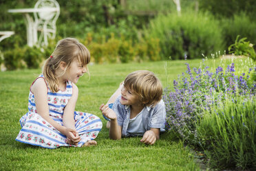 Ein Mädchen sitzt im Gras und unterhält sich mit ihrem Bruder, der neben ihr auf einer Wiese in einem Garten liegt. - MINF04419