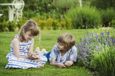 Ein Mädchen sitzt im Gras und unterhält sich mit ihrem Bruder, der neben ihr auf einer Wiese in einem Garten liegt. - MINF04420