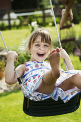 Lächelndes Mädchen im Sommerkleid auf einer Schaukel in einem Garten. - MINF04423