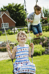 Lächelnder Junge in Hemd und Jeansshorts und Mädchen im Sommerkleid auf einer Schaukel im Garten. - MINF04424