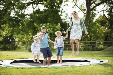 Ein Mann, eine Frau, ein Junge und ein Mädchen halten sich an den Händen und springen auf einem in den Boden eingelassenen Trampolin in einem Garten. - MINF04432