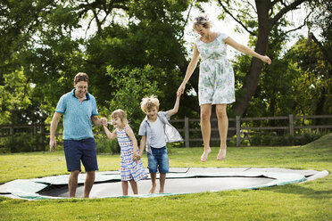 Man, woman, boy and girl holding hands, jumping on a trampoline set in the lawn in a garden. - MINF04433