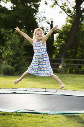Mädchen im Sommerkleid springt auf einem in den Boden eingelassenen Trampolin, in einem Garten. - MINF04434