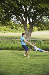 Man standing on a lawn in a garden, holding a boy by his hands, spinning and whirling him around. - MINF04443