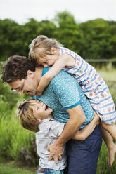 Ein Vater und zwei Kinder in einem Garten, ein Junge umarmt ihn um die Taille und ein Mädchen liegt auf seinem Rücken und hat die Arme um seinen Hals gelegt. - MINF04445
