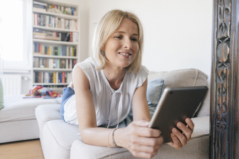 Smiling mature woman lying on couch at home using tablet stock photo