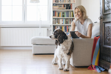 Smiling mature woman with dog lying on couch at home holding tablet - JOSF02450