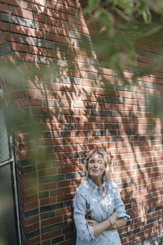 Entspannte reife Frau lehnt an einer Hauswand und hält eine Gartenschere, lizenzfreies Stockfoto