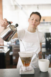 Frau mit weißer Schürze steht in einem Café und macht Filterkaffee. - MINF04611