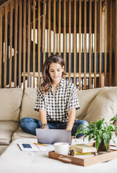 Woman with long brown hair sitting on a sofa with a laptop computer and notebook, working. - MINF04626